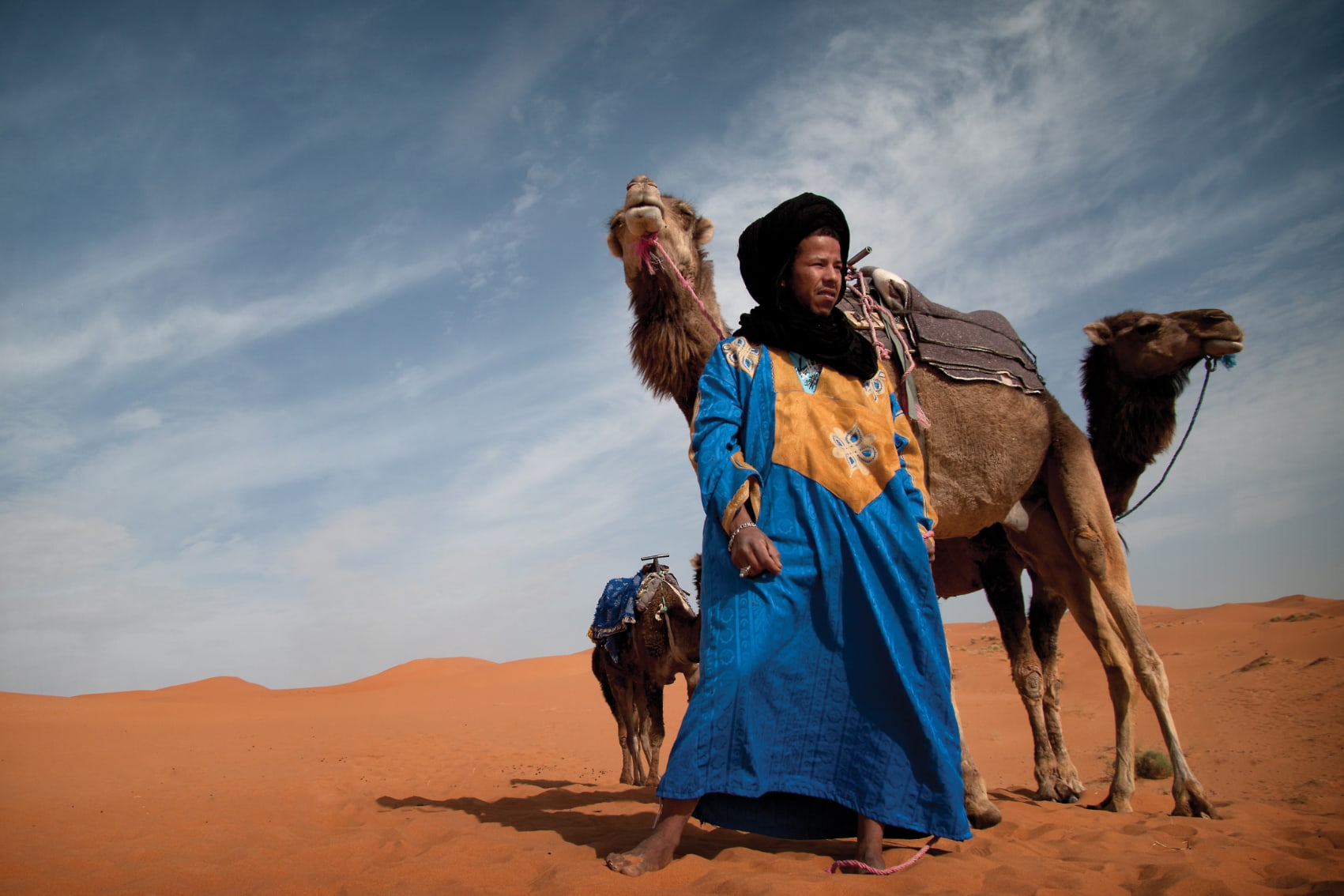 a traditionally dressed guide leads a camel through the Moroccan sand dunes